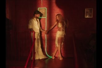 Maluma and Shakira standing side by side on a bright set, both singing into microphones with water effects surrounding them in the official AGUA music video
