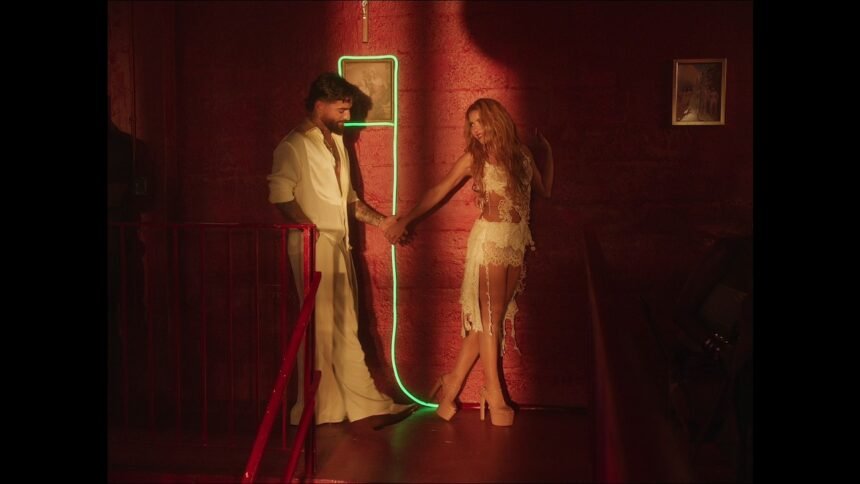 Maluma and Shakira standing side by side on a bright set, both singing into microphones with water effects surrounding them in the official AGUA music video