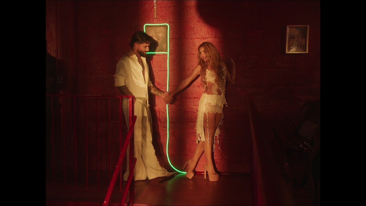Maluma and Shakira standing side by side on a bright set, both singing into microphones with water effects surrounding them in the official AGUA music video