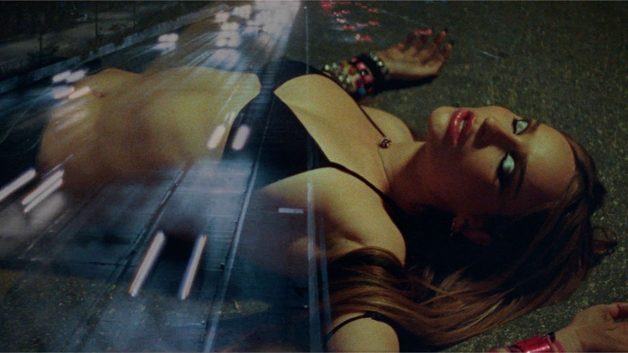 Kim Petras standing on a highway at night, wearing a dark outfit, with city lights in the background, as seen in the official 'Autobahn' music video.