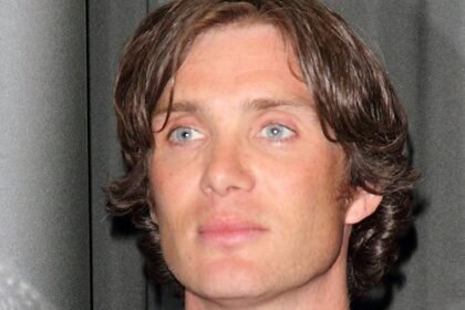 Cillian Murphy wearing a dark suit, looking serious, standing on a film set with lighting equipment behind him.