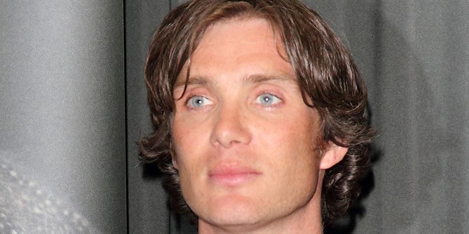 Cillian Murphy wearing a dark suit, looking serious, standing on a film set with lighting equipment behind him.