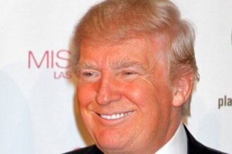 Donald Trump speaking at a political rally, wearing a dark suit and tie, with an American flag in the background.