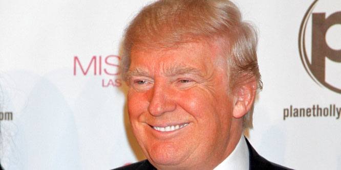 Donald Trump speaking at a political rally, wearing a dark suit and tie, with an American flag in the background.