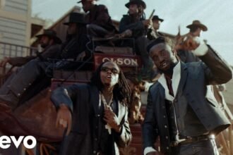 Shaboozey standing in a desert landscape with the sun overhead, wearing a black jacket, while Gunna stands beside him, both looking toward the camera, captured in the official “High Noon” video.