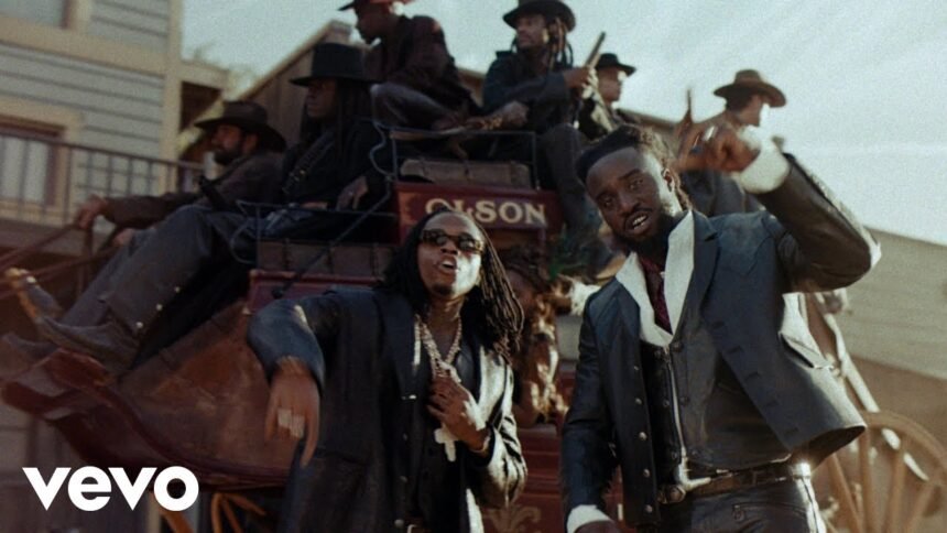 Shaboozey standing in a desert landscape with the sun overhead, wearing a black jacket, while Gunna stands beside him, both looking toward the camera, captured in the official “High Noon” video.