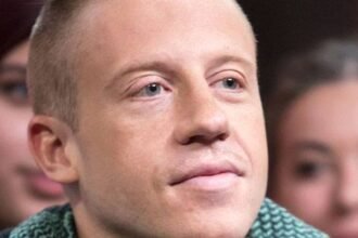 Macklemore on stage wearing a jacket, holding a microphone, with a crowd behind him