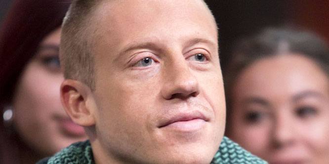 Macklemore on stage wearing a jacket, holding a microphone, with a crowd behind him