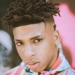 NLE Choppa on stage during a concert, wearing a black hoodie and a gold chain, with bright lights behind him.