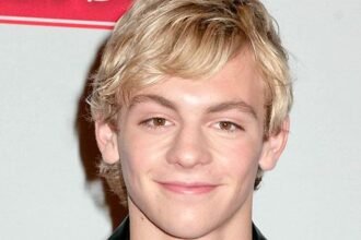 Ross Lynch sings and plays guitar on stage with his band The Driver Era, wearing a black jacket and smiling at the audience.