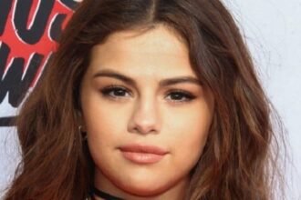 Selena Gomez smiling on a red carpet wearing a sleek black dress, with bright lights behind her.