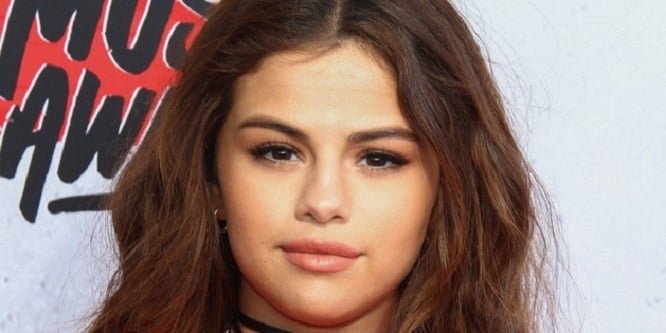 Selena Gomez smiling on a red carpet wearing a sleek black dress, with bright lights behind her.