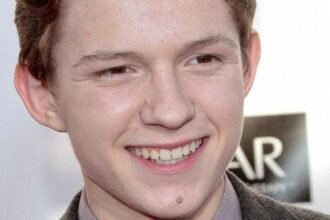 Tom Holland smiling in a suit at a movie premiere, with a backdrop of a city skyline