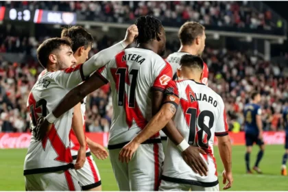 Rayo Vallecano players in red shirts and Elche players in white shirts standing on the pitch before the match at the Vallecas stadium.