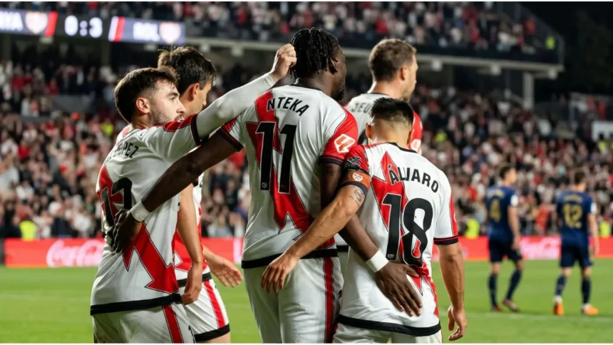 Rayo Vallecano players in red shirts and Elche players in white shirts standing on the pitch before the match at the Vallecas stadium.