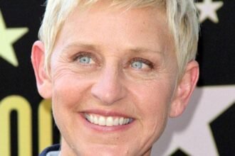 Ellen DeGeneres smiling on a red carpet, wearing a sleek black dress and holding a microphone during a talk‑show event.