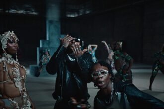 Blaqbonez and Asake performing together on a bright set with dancers in carnival outfits for the Ikebe 3000 music video