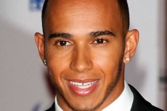 Lewis Hamilton wearing a Mercedes racing suit, smiling on the podium with a trophy and British flag in the background