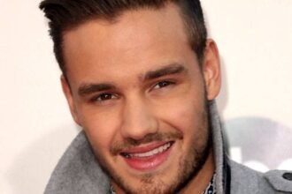 Liam Payne standing on a concert stage wearing a black jacket, holding a microphone, with bright lights behind him.