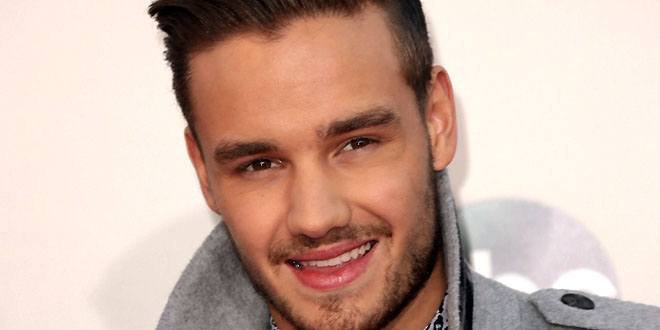 Liam Payne standing on a concert stage wearing a black jacket, holding a microphone, with bright lights behind him.