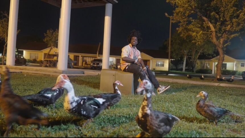 Kodak Black standing in a dimly lit studio, wearing a hoodie, with the title "Little Birdie" displayed on screen behind him