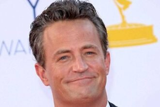 Matthew Perry smiling while filming the TV show Friends, wearing a casual shirt and holding a coffee mug.