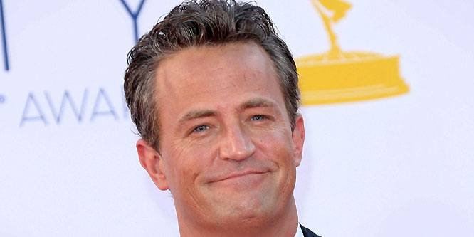 Matthew Perry smiling while filming the TV show Friends, wearing a casual shirt and holding a coffee mug.