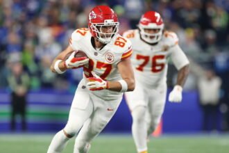 Travis Kelce in a Kansas City Chiefs uniform leaping to catch a football, with teammates and fans in the background