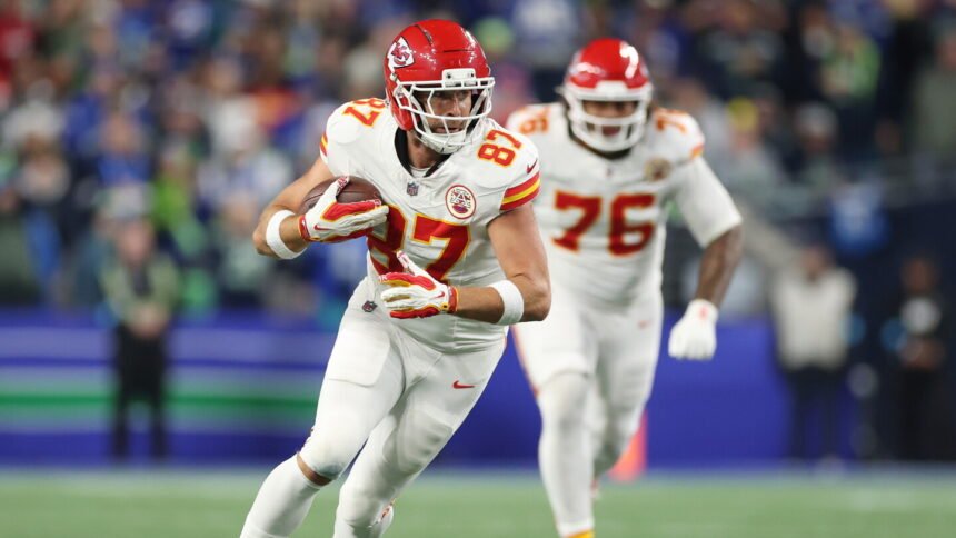 Travis Kelce in a Kansas City Chiefs uniform leaping to catch a football, with teammates and fans in the background